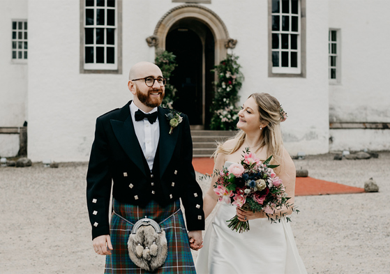 A bride and groom walk hand in hand outside a large white Scottish Castle after their wedding ceremony.